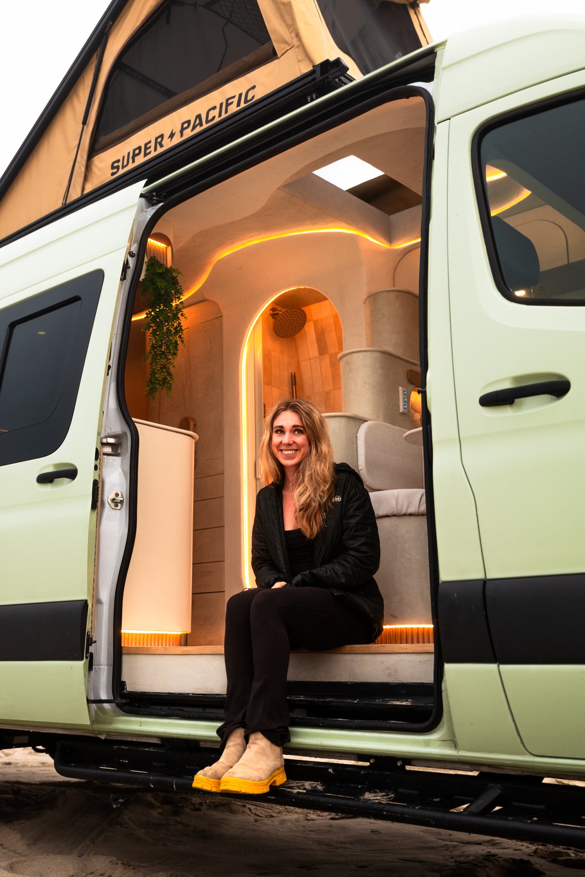 Abbe sitting in Flow's open slider door on the sand, the Super Pacific tent popped and the plaster interior lit amber behind her